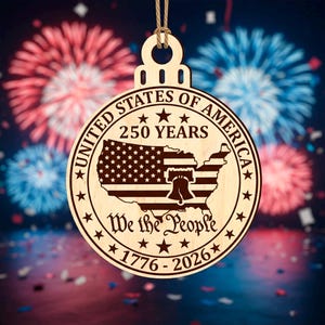 Peut inclure: Ornement en bois de forme circulaire avec un design patriotique. Il porte l'inscription "UNITED STATES OF AMERICA 250 YEARS" et "We the People 1776-2026". Le design comprend une carte des &Eacute;tats-Unis avec le drapeau am&eacute;ricain et la Cloche de la Libert&eacute;.