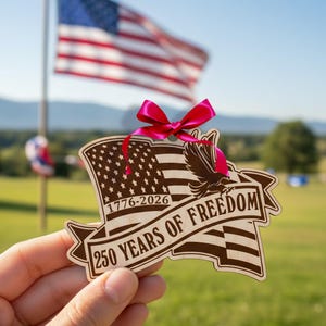 Peut inclure: Ornement en bois avec un drapeau am&eacute;ricain, le texte indique "1776-2026" et "250 YEARS OF FREEDOM." Il est orn&eacute; d'un ruban rose. Un drapeau am&eacute;ricain flotte en arri&egrave;re-plan.