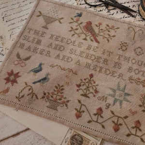 May include: A cross-stitch sampler with a beige background and brown, red, and blue embroidery. The sampler features a quote: "The needle be it though so slim and slender is both a maker and a mender."