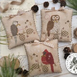 May include: Three cross-stitch patterns featuring a cardinal, an owl, and a bird on a branch. The patterns are stitched on beige fabric and are framed with a white border. The patterns are titled "Peppermint & Pine" and are designed by Brenda Gervais.