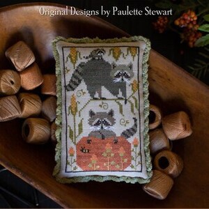 May include: A cross-stitch sampler featuring three raccoons, one sitting on a pumpkin, the others perched on a branch. The sampler is framed in a green fabric with a scalloped edge and is displayed in a wooden bowl with wooden beads. The title "Raccoon Rabble" is displayed below the sampler.