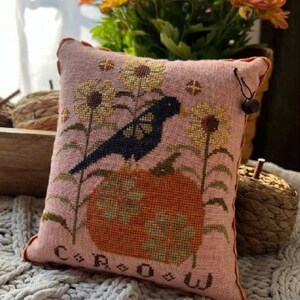 May include: A pink cross-stitch pillow with a black bird, a pumpkin, and flowers. The pillow reads "CROW" and is designed by Annie Belcher. The pillow is on a white fuzzy surface with a bouquet of orange flowers in the background.