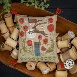 May include: A cross-stitch design featuring a rabbit carrying an American flag, surrounded by red poppies and a blue house. The design is on a small pillow, displayed in a wooden bowl with spools of thread. The text "Plum Street Samplers" is at the top.