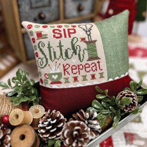 May include: A cross-stitch pillow with a red, green, and white design. The pillow reads "Sip Stitch Repeat" and features a sewing needle, thread, and scissors. The pillow is sitting on a wooden table with a red and green Christmas tree in the background.