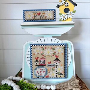 May include: A cross-stitch pattern featuring a birdhouse, birdseed, and a clock with the words "Bird Seed" and "Home". The pattern is framed in blue and white and is displayed on a vintage scale with a small cross-stitch sign that says "May".