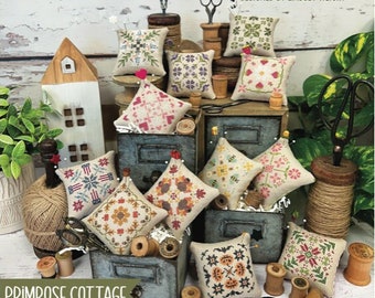 May include: A collection of small, square, embroidered pillows with various floral and geometric designs. The pillows are arranged on and around vintage-style wooden boxes, with spools of thread, scissors, and the text "12 MONTHLY MINIS" and "PRIMROSE COTTAGE".