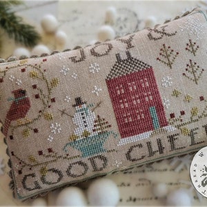 May include: A cross-stitch pillow with a winter scene featuring a red house, a snowman, and cardinals. The pillow is beige with brown stitching and the text "Joy & Good Cheer" is stitched on the pillow.