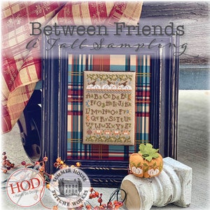 May include: A framed cross-stitch sampler with the text "Between Friends A Fall Sampling" and the alphabet in a vintage style. The sampler is in a blue frame and is sitting on a plaid fabric with a small pumpkin ornament.
