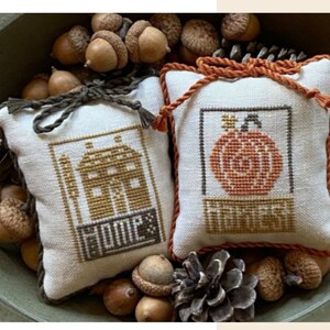 May include: Two small white pillow-shaped ornaments with embroidered designs. One features a house with the word "Home" and the other has a pumpkin with the word "Harvest". The ornaments are surrounded by acorns and pine cones in a brown bowl.