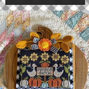 May include: A cross-stitch pattern titled Pumpkin Patch Geese featuring two white geese, orange pumpkins, and yellow sunflowers on a dark fabric background. The piece is accented with a crocheted yellow flower, a black and white gingham bow, and leaves.