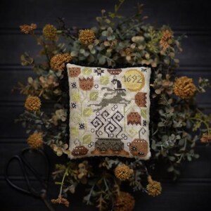 May include: A cross-stitch design titled "Salem Mini Moon" featuring a witch, pumpkins, and the year 1692. The design is set against a backdrop of greenery and dried flowers, with the "Plum Street Samplers" logo at the top.