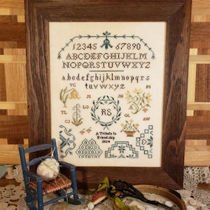 May include: A framed cross-stitch sampler with a white background and colorful floral designs. The sampler includes the alphabet, numbers, and the initials "RS". The text "A Tribute to Friendship 2024" is also included.
