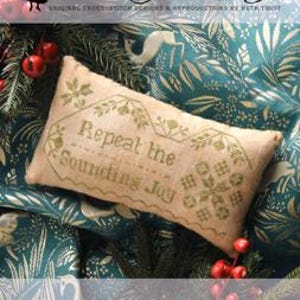 May include: A rectangular, beige cross-stitch pillow with the text "Repeat the Sounding Joy" in green thread. The pillow is decorated with floral and geometric patterns. The background is a teal fabric with a botanical print, and red ornaments are scattered around.