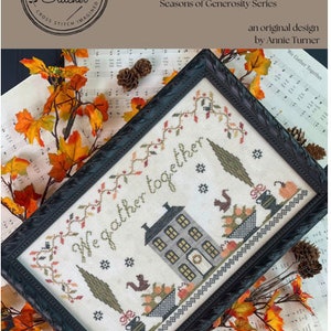 May include: A cross stitch pattern featuring a house with a wreath on the door, two trees, and a basket of pumpkins. The text "We gather together" is stitched above the house. The pattern is framed in a black frame and surrounded by fall leaves and pine cones.