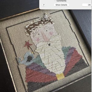May include: A framed cross-stitch embroidery of a cartoon character with a crown, wearing a colorful patterned shirt and holding a star. The character has a blue bird on its shoulder.