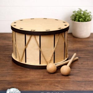 May include: A wooden drum with a light brown finish, black trim, and rope detailing. Two wooden drumsticks rest beside the drum. The word "DRUM" is printed in white on a blue banner.