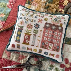 May include: A square, off-white pillow with a cross-stitched design featuring a pink house, spools, and flowers. The text "button house" is stitched at the top. The pillow has a teal scalloped edge. A pair of decorative scissors rests below.