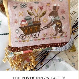 May include: A decorative pillow with a cross-stitch design. The pillow features a brown bunny pushing a cart filled with Easter eggs and flowers. The text "THE POSTBUNNY'S EASTER DELIVERY" is at the bottom. The pillow has a pink background with a yellow border.