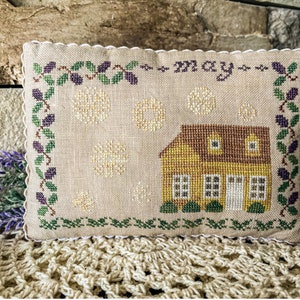 May include: A beige pillow with a scalloped edge features a cross-stitch design of a yellow house with a brown roof and green trim. The house is surrounded by white flowers and green leaves. The text "may" is stitched in the top center of the pillow.