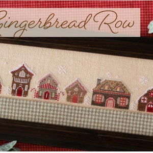 May include: A framed cross-stitch design featuring a row of gingerbread houses with candy canes and snow. The title "Gingerbread Row" is displayed above the houses.