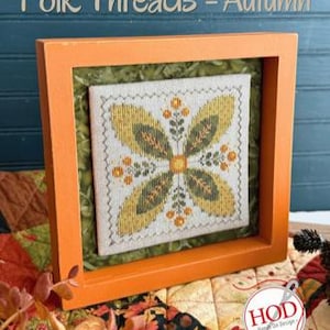 May include: A square cross-stitch embroidery featuring a symmetrical floral motif in shades of yellow, olive green, and orange on a white fabric background, framed in a bright orange wooden shadow box with the text Folk Threads - Autumn.