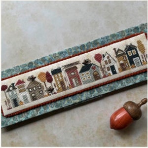 May include: Cross-stitch pattern of a row of houses with autumnal colors and details, including trees, a bird, and a hot air balloon. The houses are in various styles and colors, with some having chimneys and windows. The pattern is framed with a blue and green border.
