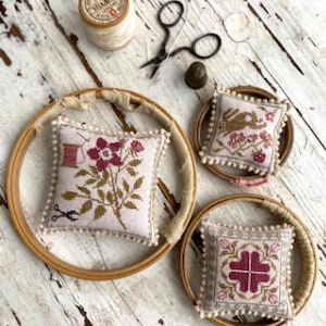 May include: Three embroidery hoops with different designs. The first hoop has a floral design with pink flowers and green leaves. The second hoop has a design of a rabbit with pink flowers and green leaves. The third hoop has a geometric design with a pink and white pattern.