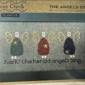 May include: A framed cross-stitch design featuring three angels with wings and stars. The text "hark! the herald angels sing." is stitched below the angels.