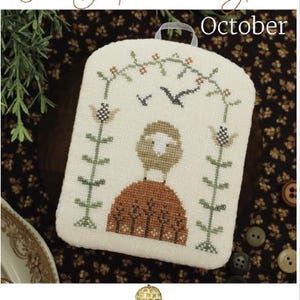 May include: A cross-stitch design featuring a sheep with a brown sweater and a floral arch. The text "The Shepherd's Year" and "October" are displayed. The design is on a cream-colored fabric with a small handle.