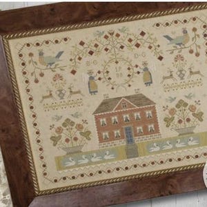 May include: Cross-stitch sampler titled "Manor at Swan Lake" by Brenda Gervais, framed in a dark wood frame. The sampler features a house, birds, deer, and floral elements in shades of red, blue, and green. The text "Manor at Swan Lake" is visible.
