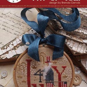 May include: A cross-stitch embroidery hoop with a patriotic 4th of July theme. The design features the words "4th of July" in red, a depiction of a figure in red, white, and blue, and a blue ribbon for hanging.