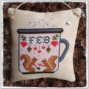 May include: A cross-stitched pillow with a coffee mug design. The mug features the word "FEB", hearts, and squirrels. The pillow is beige with a jute hanger and a small heart charm.