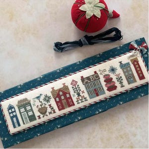 May include: A cross-stitch bookmark featuring a row of colorful houses, trees, and flowers. The bookmark is bordered with a red and white twisted cord. A red strawberry pin cushion, embroidery floss, and small scissors are also visible.