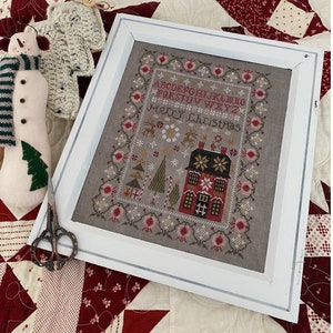 May include: A framed cross-stitch design featuring a winter scene with a house, trees, and the text "Merry Christmas". The alphabet is also stitched in the design.