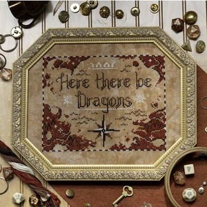 May include: Framed cross-stitch art with the text "Here there be Dragons" in a gold-colored frame. The artwork features a map design with red and brown accents. Various crafting supplies, including dice, buttons, and thread, surround the frame.