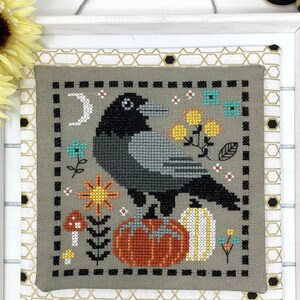 May include: Cross stitch pattern featuring a black crow perched on a pumpkin with a crescent moon and floral accents. The design is framed in a gray fabric with a black border.