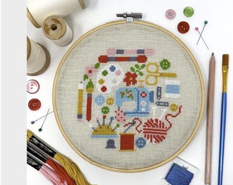 May include: A cross-stitch embroidery hoop with a colorful design featuring sewing and art supplies. The design includes a sewing machine, paint palette, scissors, and yarn. Additional supplies include pencils, a paintbrush, and spools of thread.