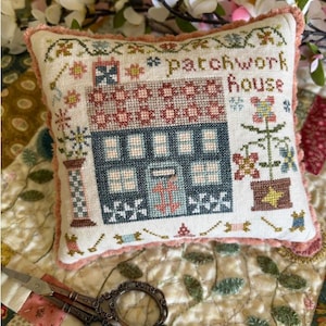 May include: A decorative, square, cross-stitched pillow with the words "patchwork house". The design features a blue house with a red roof, surrounded by floral and geometric patterns. The pillow has a pink fringed border and is set on a floral quilt.