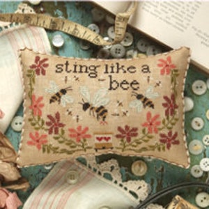 May include: A rectangular, beige cross-stitch pillow with the text "STING LIKE A BEE" and images of bees and flowers. The pillow is surrounded by sewing notions, including buttons, a measuring tape, and lace.
