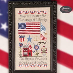 May include: A framed cross-stitch sampler with patriotic imagery. The design includes an American flag, an eagle, and the text "May we secure the Blessings of Liberty." The sampler also features a red house, flowers, and the text "The Hope & Promise of the Land of the Free."