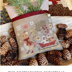 May include: A beige cross-stitched pillow with a mouse pulling a sleigh filled with gifts. The pillow is decorated with snowflakes and greenery, and is surrounded by pine cones. The text "THE POSTMOUSE'S CHRISTMAS DELIVERY" is at the bottom.