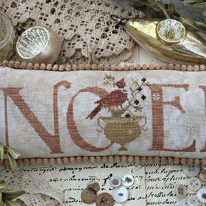 May include: A rectangular, off-white pillow with the word "NOEL" embroidered in a rust-colored thread. A bird and floral arrangement are in the center. The pillow is trimmed with a light brown pom-pom border. Decorative Christmas ornaments are in the background.
