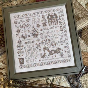 May include: A framed cross-stitch sampler on off-white fabric featuring brown and muted-tone embroidery of squirrels, a house, flowers, and the alphabet in both uppercase and lowercase letters, along with the numbers 1 through 10 in a gray frame.