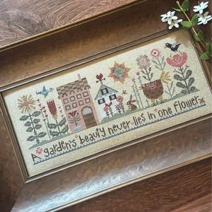 May include: A framed cross-stitch sampler featuring a quaint village scene with houses, flowers, and a bird. The text "A garden's beauty never lies in one flower" is stitched below the scene. The frame is made of wood.