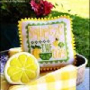 May include: A cross stitch pattern with a yellow lemon and the text "Squeeze the Day" in a green and yellow font. The pattern is on a white background with a pink border. The pattern is framed and displayed on a yellow and white checkered surface.