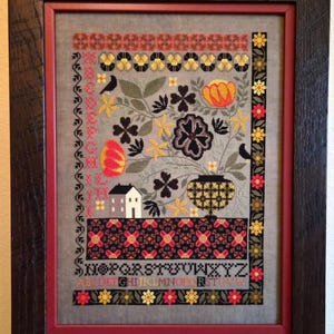 May include: Framed cross-stitch sampler with a gray background. The design features floral and geometric patterns in red, yellow, black, and green. A house and birds are also included. The frame is dark brown with a red inner border.