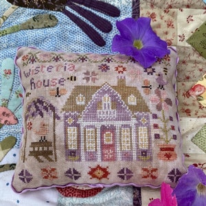 May include: A light purple and brown cross-stitch pillow with the text "wisteria house" on the front. The pillow is decorated with a small house, flowers, and a bird.
