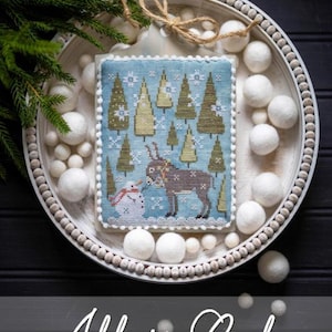 May include: A cross-stitch Christmas ornament with a winter scene featuring a reindeer and snowman. The ornament is surrounded by white pom-poms and greenery. The text "All is Calm" is at the bottom.
