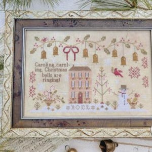 May include: Framed cross-stitch artwork with a Christmas theme. The design features a house, snowman, and bells, with the text "Caroling, caroling, Christmas bells are ringing!". The frame is ornate with a light-colored finish.