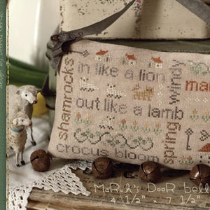 May include: A cross-stitch sampler with the text "shamrocks in like a lion march out like a lamb crocus bloom" and "MoRak's Door bells" in a brown frame. The sampler is surrounded by small bells and a book.